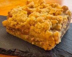 A slice of crumble cake with a golden topping on a slate plate.